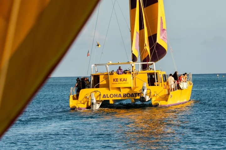 Yellow catamaran with colorful sails on the ocean, viewed from another boat.