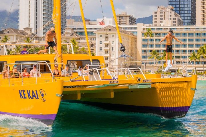 People enjoying a sunny day on a yellow catamaran near city buildings.